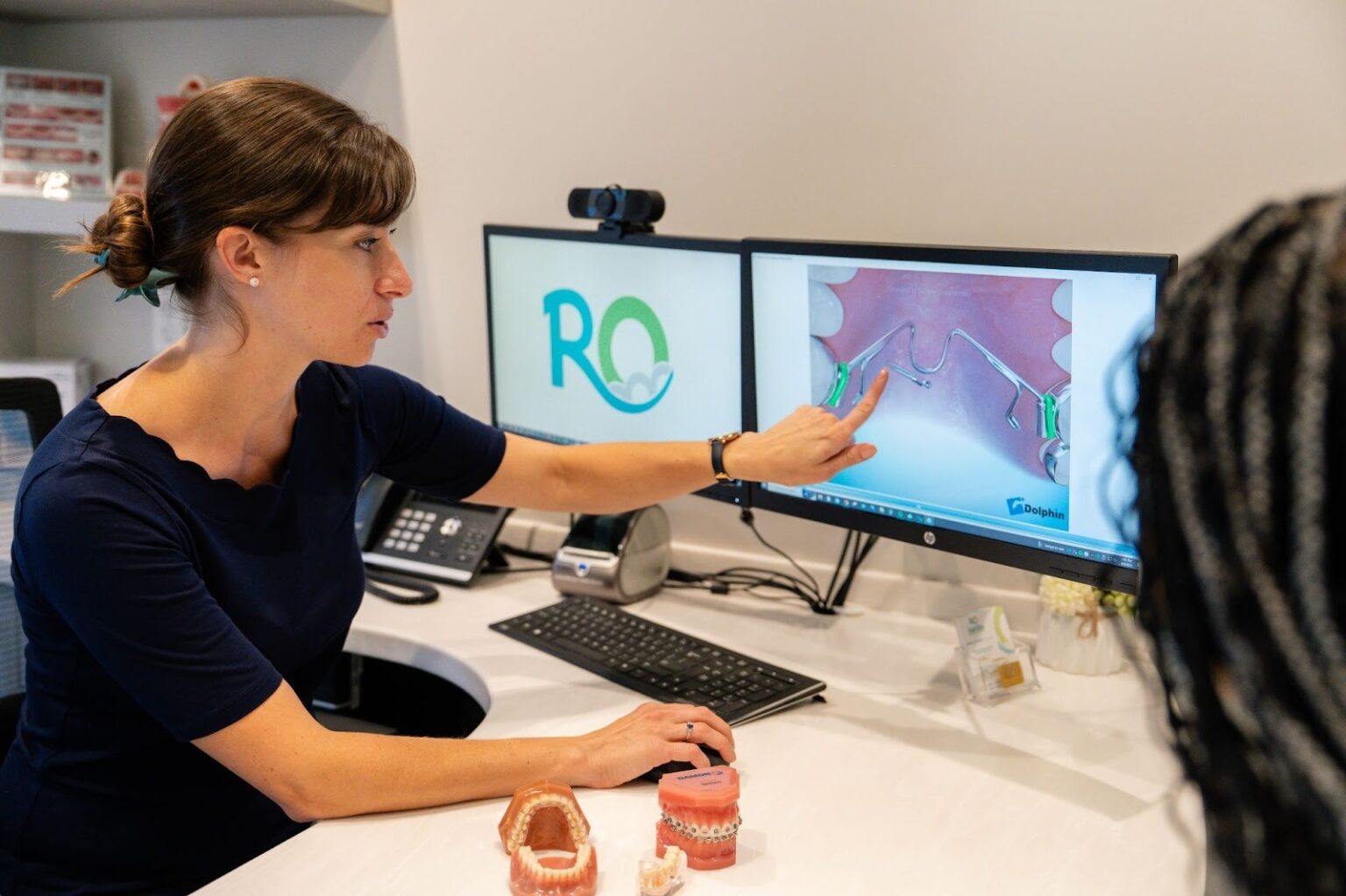 Orthodontist explaining treatment plan using digital imaging on computer screens, featuring dental models on desk, in a modern orthodontic office setting.